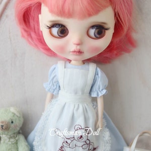May include: A doll with pink hair wearing a light blue dress with a white apron and a brown embroidered bear design. The doll is holding a green teddy bear and a small brown and red bag with a bear design.