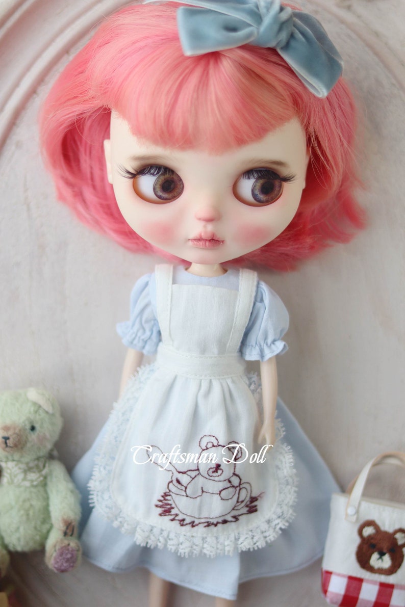 May include: A doll with pink hair wearing a light blue dress with a white apron and a brown embroidered bear design. The doll is holding a green teddy bear and a small brown and red bag with a bear design.