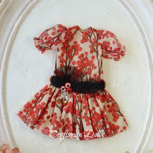 May include: A miniature doll dress with a beige background and a red berry pattern. The dress features short, puffed sleeves, a black lace belt, and decorative embellishments. The text "Craftsman Doll" is visible.