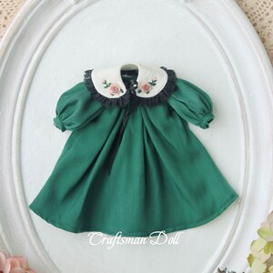 May include: A green dress with a white collar and black lace trim. The collar is embroidered with pink roses. The dress has a black ribbon tie at the neck. "Craftsman Doll" is printed on the bottom of the dress.
