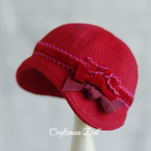 Könnte beinhalten: Ein roter Filzhut mit einem rosa und burgunderfarbenen Band und Schleife. Der Hut steht auf einem weißen Ständer.
