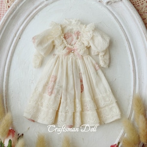 May include: A white dress with pink floral print and lace trim. The dress is displayed on a white oval frame with a white background. The text "Craftsman Doll" is visible at the bottom of the image.