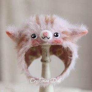 Puede incluir: Un gorro de gato de color rosa y marrón peludo con ojos negros y una nariz blanca. El gorro está en un soporte blanco y el texto "Craftsman Doll" es visible debajo del gorro.