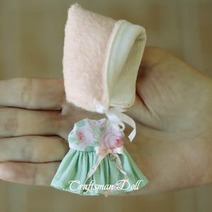 May include: A miniature doll dress with a green skirt and a floral print bodice. The dress is paired with a pink bonnet.