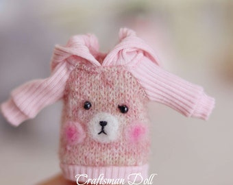 May include: A pink knitted sweater with a bear face design and a hood. The sweater has a ribbed texture and is made for a small doll.