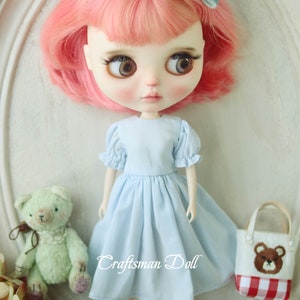 May include: A doll with pink hair wearing a light blue dress and a blue bow. The doll is holding a small green teddy bear and a red and white checkered bag with a brown bear on it. The text "Craftsman Doll" is visible on the image.