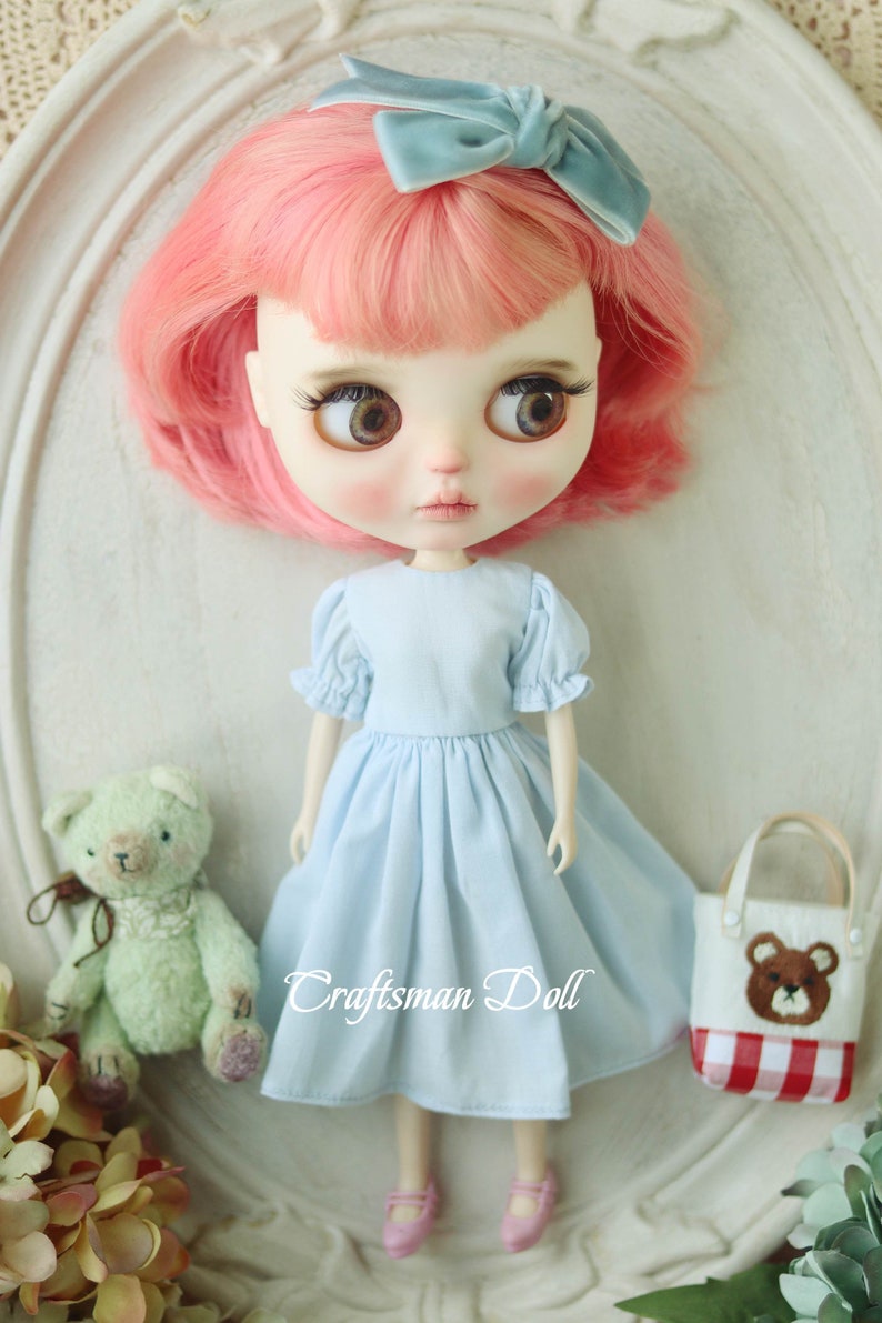 May include: A doll with pink hair wearing a light blue dress and a blue bow. The doll is holding a small green teddy bear and a red and white checkered bag with a brown bear on it. The text "Craftsman Doll" is visible on the image.