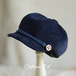 Könnte beinhalten: Eine marineblaue Cord-Newsboy-Mütze mit blauem Seilbesatz und einem goldenen Knopf mit einem weißen Blumenmuster.