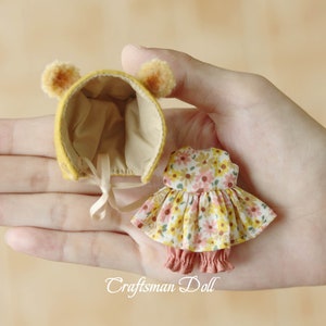 Könnte beinhalten: Ein Miniatur-Puppen-Outfit mit einer gelben Mütze mit Bärenohren und einem Blumenkleid mit passenden Bloomers. Das Kleid hat ein gelbes und rosa Blumenmuster. Der Text "Craftsman Doll" ist sichtbar.