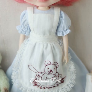 May include: A light blue doll dress with a white apron featuring a brown embroidered teddy bear design. The apron has a lace trim and the dress has puffed sleeves.