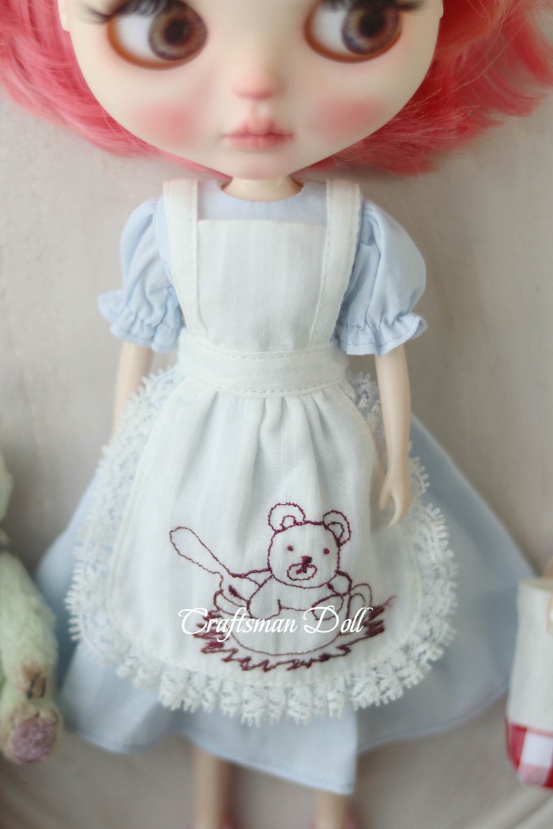 May include: A light blue doll dress with a white apron featuring a brown embroidered teddy bear design. The apron has a lace trim and the dress has puffed sleeves.