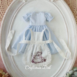 May include: A white doll apron with a brown embroidered teddy bear design and lace trim. The apron is paired with a light blue dress with puffed sleeves.