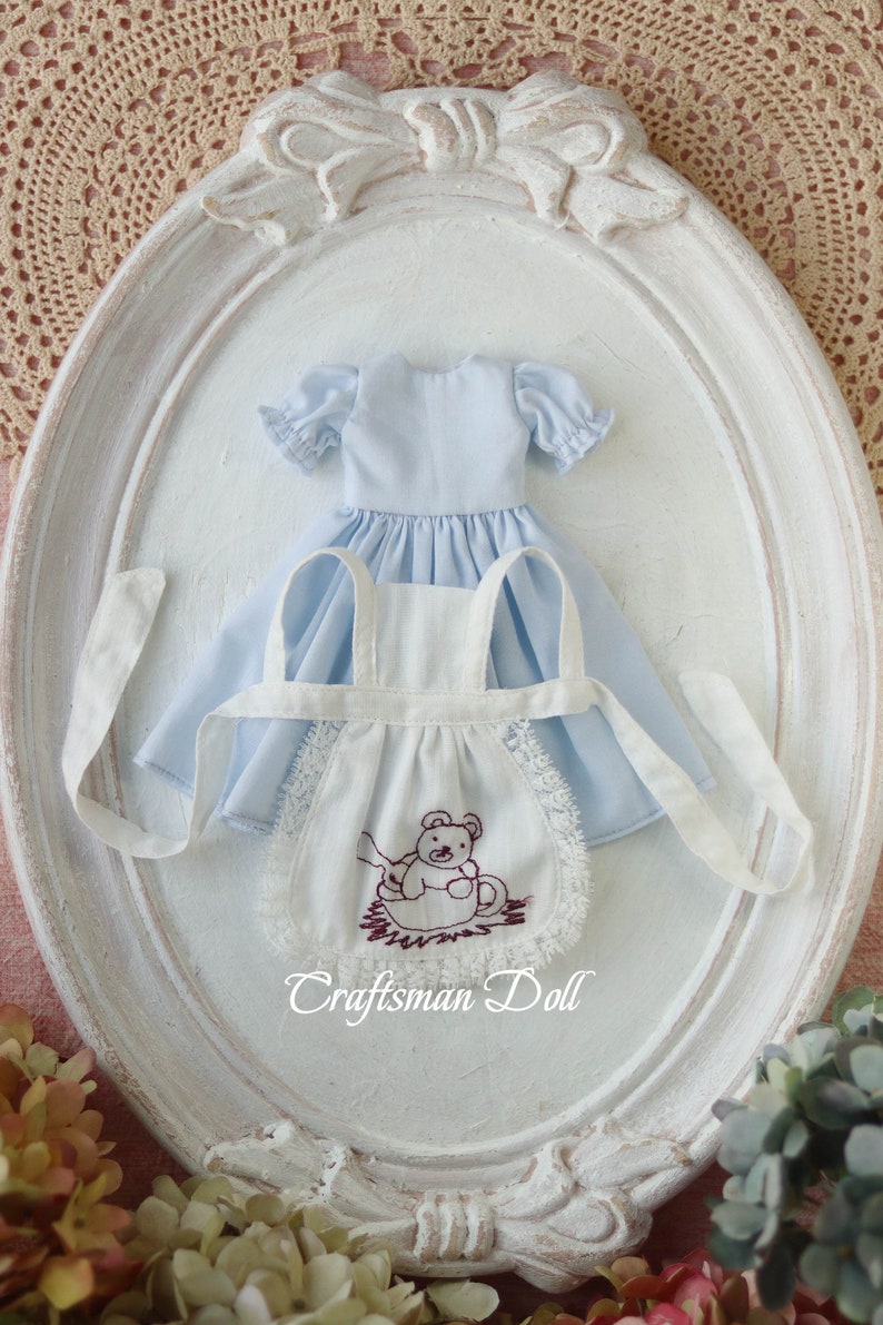 May include: A white doll apron with a brown embroidered teddy bear design and lace trim. The apron is paired with a light blue dress with puffed sleeves.