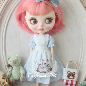 May include: A doll with pink hair wearing a light blue dress with a white apron and a brown teddy bear embroidered on it. The doll is holding a small white bag with a red and white checkered pattern and a brown teddy bear embroidered on it. The doll is standing on a white surface with a lace pattern.