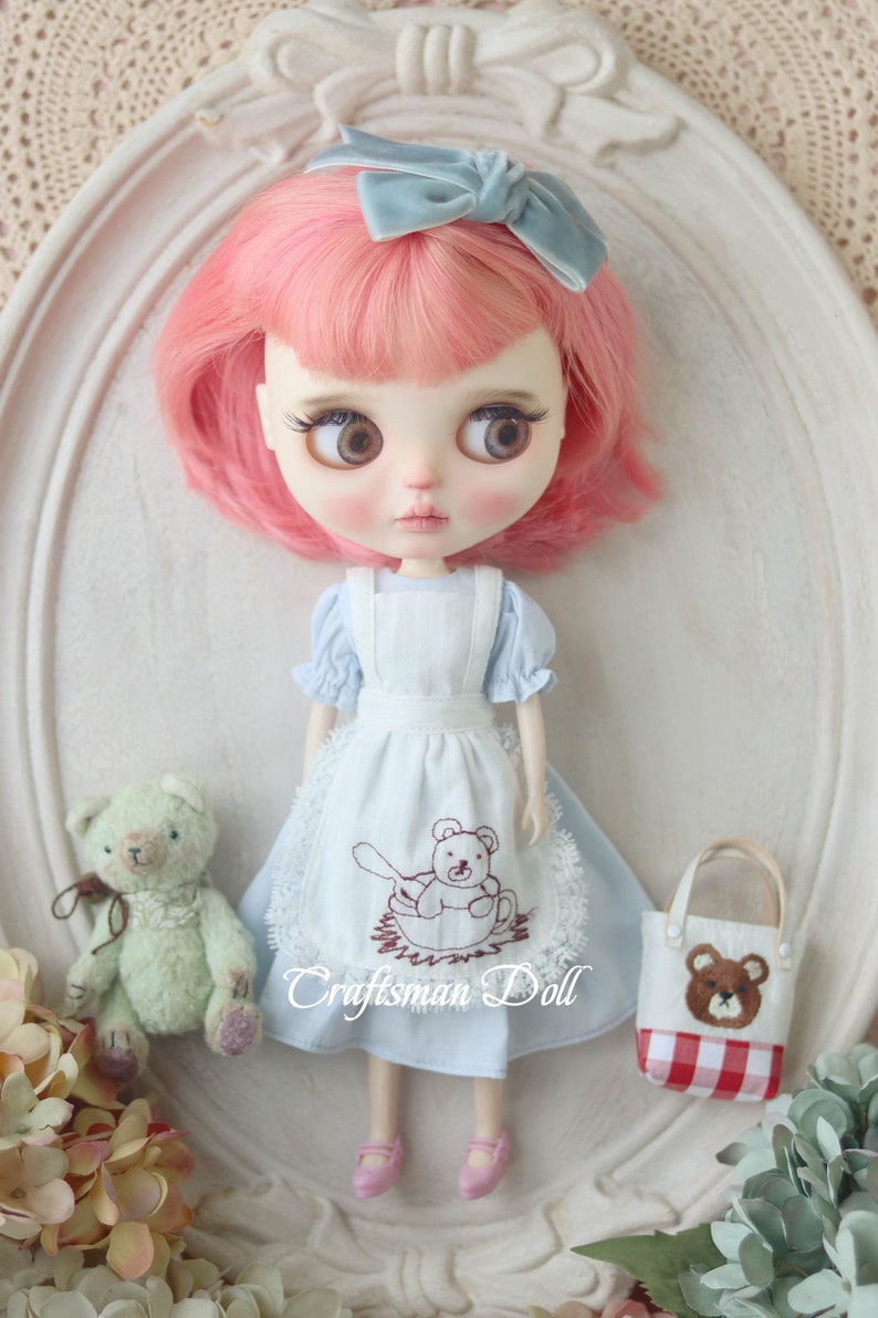 May include: A doll with pink hair wearing a light blue dress with a white apron and a brown teddy bear embroidered on it. The doll is holding a small white bag with a red and white checkered pattern and a brown teddy bear embroidered on it. The doll is standing on a white surface with a lace pattern.