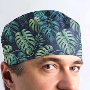 May include: A dark blue scrub hat with a tropical leaf pattern. The hat features green and white monstera leaves on a dark blue background.