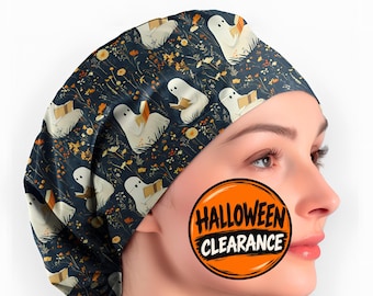 May include: A dark blue scrub cap featuring a repeating pattern of white ghosts reading books amidst small yellow and orange wildflowers. A circular orange badge with black text reads HALLOWEEN CLEARANCE in the center of the image.