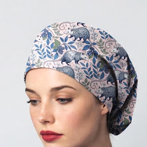 May include: A scrub cap featuring a light pink background with a repeating pattern of gray opossums, blue leaves, and dark blue berries. The fabric head covering has a rounded shape and is designed to fit snugly around the head.