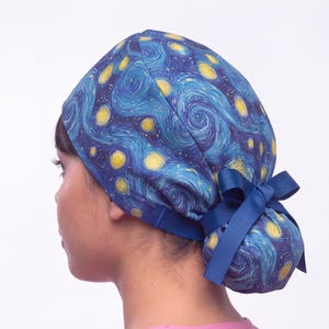May include: A scrub cap featuring a blue and yellow swirling starry night pattern. The cap has a gathered back section secured with a solid dark blue ribbon tied into a bow, providing a snug fit around the head with a smooth fabric construction.