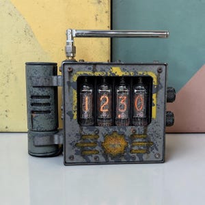May include: A rectangular gray metal clock featuring four glowing orange nixie tubes displaying the numbers 1, 2, 3, and 0. The device has a metallic antenna on top, a cylindrical side component with horizontal vents, and two black knobs on the right side.