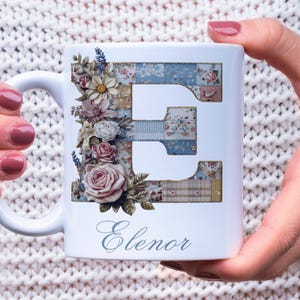 May include: White ceramic mug featuring a floral design and the letter "E" in a patchwork pattern. The mug is adorned with roses, daisies, and lavender. The name "Elenor" is written in script below the letter.