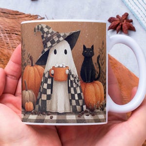 May include: White ceramic mug featuring a whimsical Halloween design. The illustration depicts a ghost wearing a checkered hat and blanket, holding a mug of cocoa. A black cat sits atop a pumpkin, with other pumpkins and coffee beans scattered around.