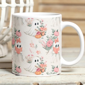 Può includere: Tazza in ceramica bianca con un motivo ripetuto di fantasmi amichevoli, zucche e composizioni floreali in rosa, arancione e verde. I fantasmi tengono libri e trasportano zucche. La tazza ha un manico bianco.