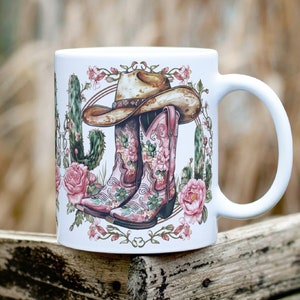 May include: White ceramic mug with a floral design featuring pink cowboy boots, a brown cowboy hat, and green cactus plants.