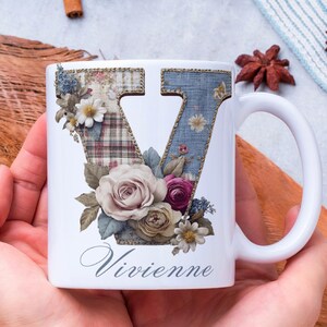 May include: White ceramic mug featuring the letter "V" decorated with floral and patchwork designs. The mug has a white handle and the name "Vivienne" in script. The floral arrangement includes roses and other flowers.