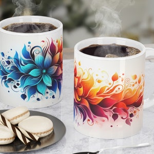 May include: Two white ceramic mugs with floral designs. One mug has a blue floral design and the other has a red, orange and yellow floral design. Both mugs are filled with coffee.