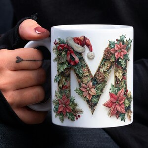 Pode incluir: Caneca de cerâmica branca com um design de letra "M" com tema natalino. A letra é feita de folhas de azevinho verdes, flores de poinsettia vermelhas e pinhas. Um chapéu de Papai Noel vermelho fica em cima da letra.