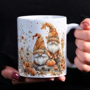 Könnte beinhalten: Weiße Keramiktasse mit einem Herbst-Motiv mit zwei Gnomen mit braunen Hüten und weißen Bärten, umgeben von Herbstlaub, Kürbissen und Blumen.