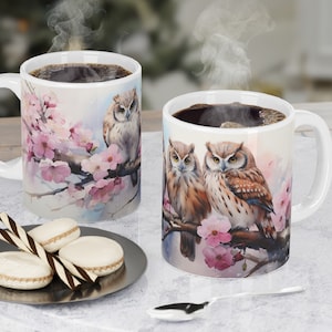May include: Two white ceramic mugs featuring a design of owls perched on a branch with pink blossoms. The mugs are filled with coffee and steam is rising from the top of the mug on the right.