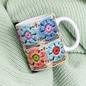 Puede incluir: Taza de cer&aacute;mica blanca con un dise&ntilde;o de flores de ganchillo de colores. La taza est&aacute; sobre una tela de punto verde claro.