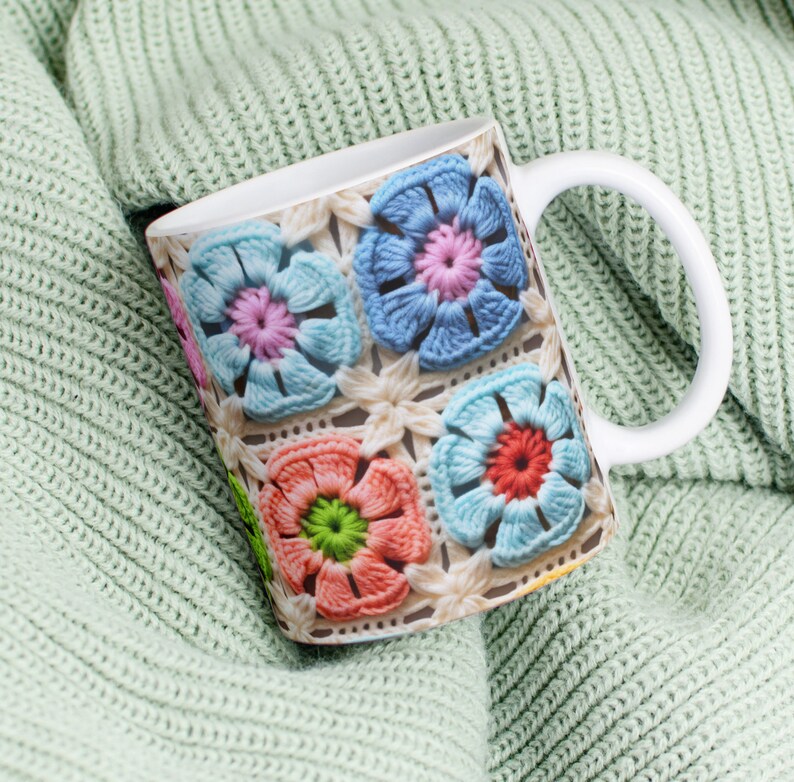 Puede incluir: Taza de cer&aacute;mica blanca con un dise&ntilde;o de flores de ganchillo de colores. La taza est&aacute; sobre una tela de punto verde claro.