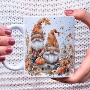 May include: White ceramic mug with a fall-themed design featuring two gnomes with orange and brown hats, surrounded by autumn leaves, pumpkins, and flowers.
