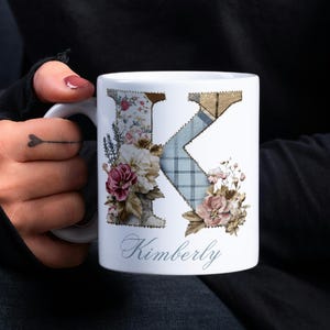 Könnte beinhalten: Weißer Keramikbecher mit dem Buchstaben "K", verziert mit Blumen- und Karomustern. Der Becher trägt den Namen "Kimberly" in Schreibschrift. Der Becher wird von einer Person gehalten.