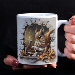 Puede incluir: Taza de cerámica blanca con una ilustración colorida de una ardilla leyendo un libro a la luz de una vela en un hueco de piedra. La ardilla es marrón y naranja con una cola tupida. El libro está abierto y tiene una cubierta amarilla. Hay otros libros y hojas de otoño en un estante debajo de la ardilla.