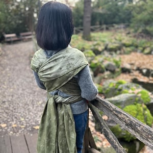 May include: A person wearing a sage green baby wrap with a floral pattern. The wrap is tied around the person's torso and shoulders. The person is wearing a grey jumper and blue jeans, standing on a wooden bridge.