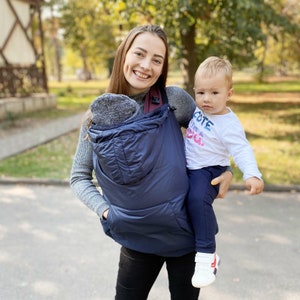 May include: A woman wearing a navy blue baby carrier with a gray hood, carrying a toddler wearing a white shirt with the text "Cute like a fox."
