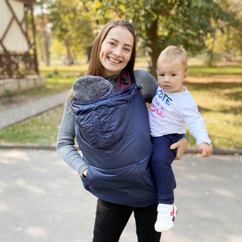 May include: A woman wearing a navy blue baby carrier with a gray hood, carrying a toddler wearing a white shirt with the text "Cute like a fox."