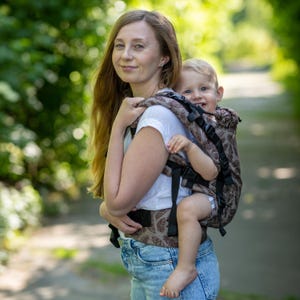 May include: A brown baby carrier featuring a repeating feather pattern in a darker shade. The carrier is constructed with black adjustable straps, plastic buckles, and a structured panel designed to support a child on the back or front.