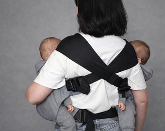 May include: A black and white baby carrier designed to hold two babies. The carrier has adjustable straps and buckles. The babies are wearing gray hooded outfits. The person wearing the carrier is wearing a white shirt and blue jeans.