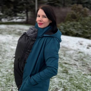 May include: Teal blue hooded jacket with a black baby carrier attached. The jacket has a high collar and a zipper closure. The person is standing outside in a snowy environment.