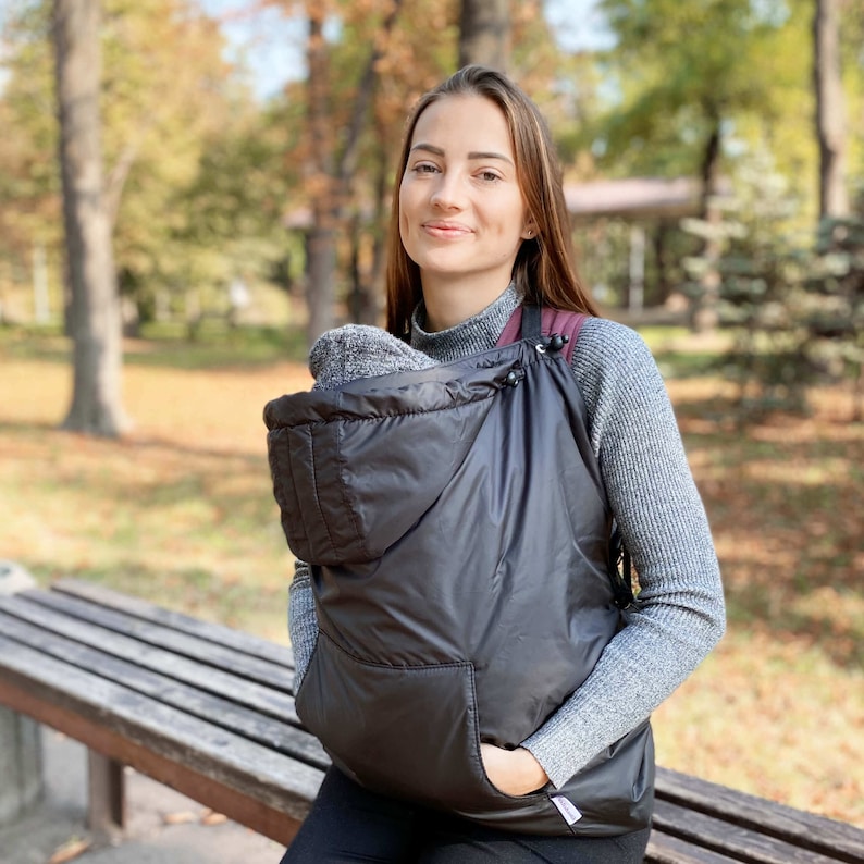 May include: A black hooded baby carrier with a zippered pocket. The carrier is worn by a person in a gray sweater and black pants. The person is standing in front of a wooden bench.