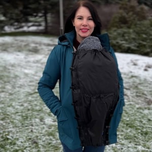 May include: A teal-colored jacket with a black baby carrier attached. The jacket has a hood and multiple pockets. The baby carrier is black and appears to be made of a water-resistant material. The background shows a snowy, outdoor setting.