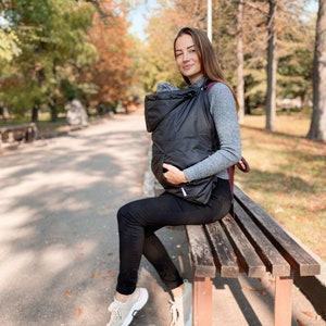 May include: A woman wearing a gray sweater sits on a wooden bench in a park. She is holding a black baby carrier with a hood. The carrier is worn over her shoulder and she is holding the carrier with one hand. The woman is looking at the camera and smiling.