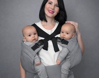 May include: A gray baby carrier designed to hold two infants. The carrier has black straps and buckles for security. The babies are wearing gray outfits. A woman is holding the babies.