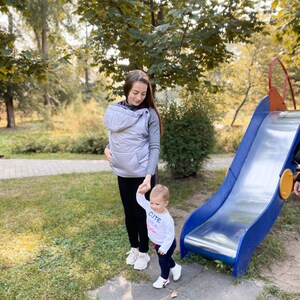 May include: A woman in a gray hooded baby carrier walks with a toddler in a white shirt that says "Cute" on a playground. The toddler is holding the woman's hand. There is a blue slide in the background.