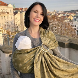 May include: An olive green baby sling with a floral design and a white metal ring. The sling is wrapped around a baby wearing a white hat. The person holding the baby is wearing a grey jumper.